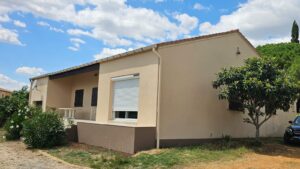 Maison beige de plain-pied avec terrasse, volets blancs et jardin fleuri sous ciel bleu.