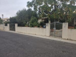 Clôture beige avec portail en fer et jardin arboré devant une maison.