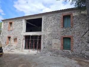Façade en pierre en rénovation avec fenêtres à briques rouges et volets verts.