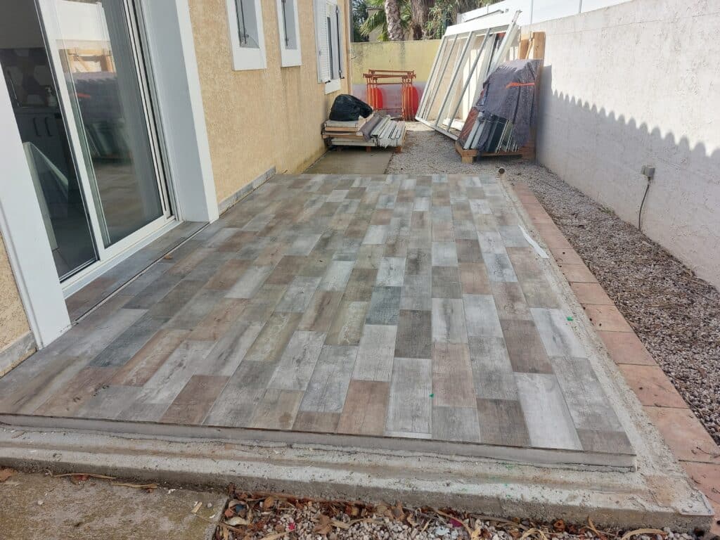 Terrasse en carrelage imitation bois gris et marron devant une porte coulissante, chantier extérieur.