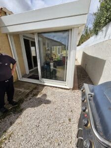 Véranda moderne avec baies vitrées sur terrasse en gravier et barbecue à gaz au premier plan.
