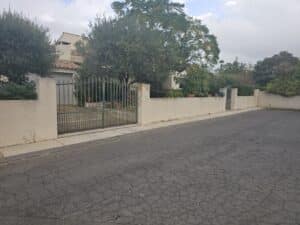 Villa, portail en fer, mur blanc et arbres devant une rue.